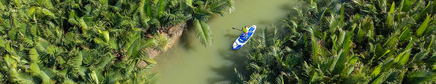 Un homme entrain de pagayer sur un stand up paddle dans une rivière à l'eau verte entre des palmiers en thailande.